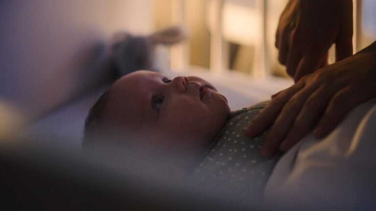 A baby lies on its back in a crib, looking up. An adult's hand is gently resting on the baby's chest, soothing during sleep regression. The setting is softly lit, creating a calm and cozy atmosphere.