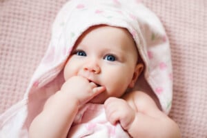 A baby wrapped in a pink towel with a hood lies on a pink textured surface, looking at the camera and sucking on their hand. The towel, adorned with small, colorful patterns, cradles the baby whose blue eyes and light skin resemble precious girl names starting with S.