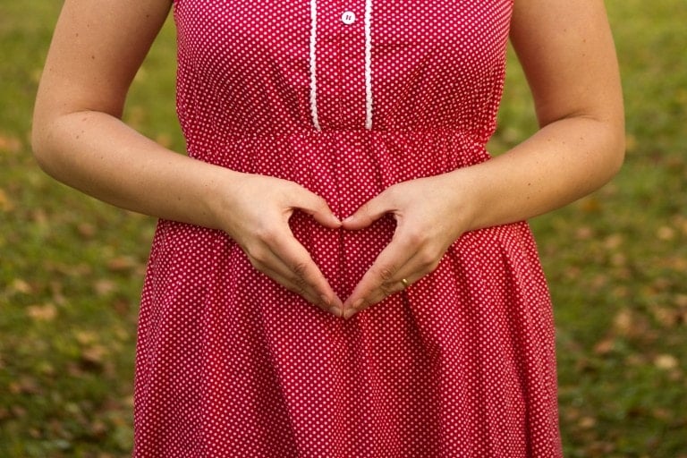 A person in a red and white polka dot dress makes a heart shape with their hands over their stomach, symbolizing the excitement of what to expect from your first trimester. The background is an out-of-focus grassy area.