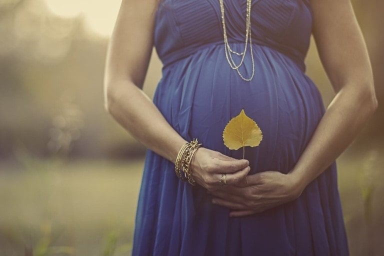 A pregnant person dressed in a blue gown holds a yellow leaf in front of their stomach, embodying perfect fall baby shower inspiration. The individual is wearing multiple bracelets on one wrist and layered necklaces. The softly blurred background suggests an enchanting outdoor setting.