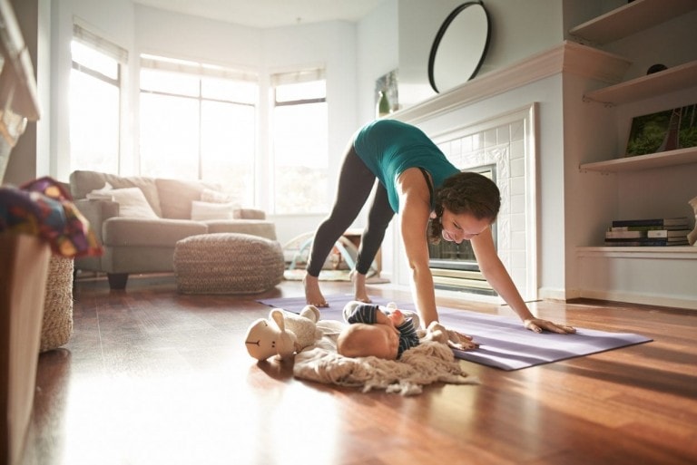 A woman is doing a yoga pose on a purple mat in a sunlit living room, showcasing how to reduce belly fat, with a baby lying on a blanket nearby and a plush toy next to the baby. The room has wooden floors, a fireplace, and large windows.