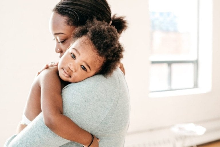 An adult is holding a young child, who rests their head on the adult's shoulder. The child looks towards the camera while the adult looks down, embodying a nurturing parenting philosophy. They are in a bright room with a window in the background.
