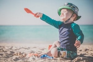 A toddler in a green and blue hat and swimwear is sitting on the sandy beach near the ocean. Holding a red plastic shovel, the child appears to be playing with beach toys, showcasing perfect Beach Essentials for Baby. The ocean and sky are visible in the background.