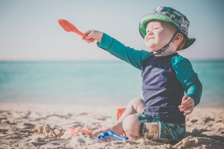 A toddler in a green and blue hat and swimwear is sitting on the sandy beach near the ocean. Holding a red plastic shovel, the child appears to be playing with beach toys, showcasing perfect Beach Essentials for Baby. The ocean and sky are visible in the background.