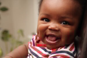 A close-up of a smiling baby boy with dark skin, wearing a red, white, and blue striped outfit. The baby has dimples and a small amount of drool is visible. The background is slightly blurred, with hints of green foliage. If you're looking for Q boy names for this adorable little one, Quinlan might fit perfectly.