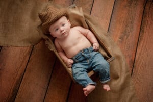 A baby wearing a brown knit cowboy hat and blue jeans lies in a wooden basket lined with burlap fabric. The basket is set on a wooden floor with burlap fabric draped around it. This scene captures the charm of country baby boy names, as the little one gazes up with a neutral expression.