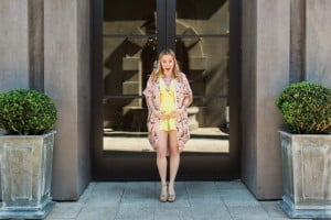 A woman in a yellow dress and floral kimono, who appears to be in the second trimester of pregnancy, stands between two potted plants in front of a large glass door. She has a surprised expression on her face with her hands placed on her stomach. The background includes a stone building facade.