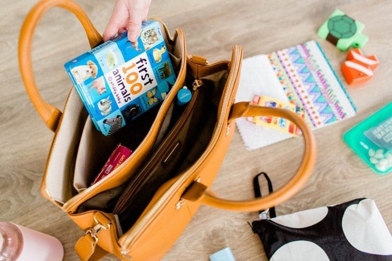 A tan toddler bag is open, revealing items inside, including a book titled "first 100 animals" and other small objects. A hand is holding the book. Nearby, other items like a patterned pouch, a block toy, and baby wipes are scattered on the wooden surface.