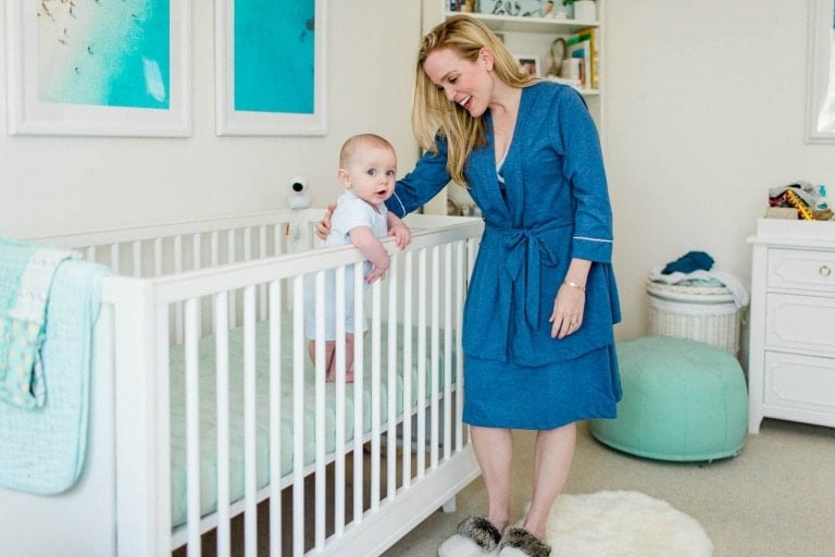 A woman, wearing a blue robe and slippers, stands next to a white crib with a baby inside. The room is decorated with blue-themed artwork and has a white dresser, a green pouf, and a round white rug on the floor. The baby is standing and holding onto the crib railing – an ideal scene for Mother's Day gifts for new moms.