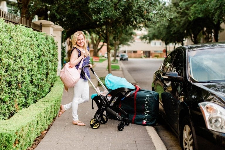 A woman with long blonde hair stands on a sidewalk next to a black car. She is wearing a blue checkered shirt and white pants, holding the handle of a baby stroller covered with a blue and black cover, showcasing her baby travel essentials. The street is lined with trees and residential buildings.