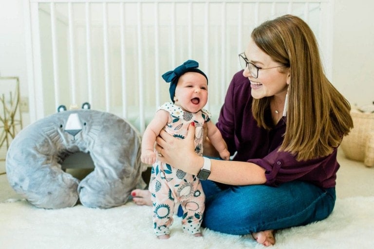 A woman with glasses and long hair, dressed in a purple top and jeans, is sitting on a rug and holding a smiling baby standing in front of her. The baby, wearing a patterned outfit and headband, looks adorable. Behind them is a white crib, gray cushion, and a Boppy pillow mentioned on her favorite safe sleep podcast.
