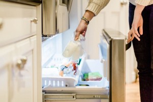 A person is seen placing or retrieving frozen breast milk from a bottom freezer drawer. The freezer contains various other items, with ice visible. The individual wears a light-colored sweater and is standing next to the open freezer in a kitchen.
