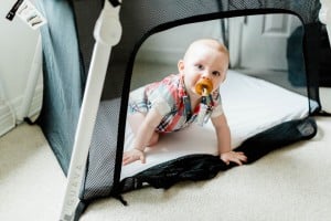 A baby is crawling out of a black and white playpen at grandma's house. The baby is wearing a plaid romper and has a pacifier in their mouth. The playpen is set on a beige carpeted floor with white walls in the background.