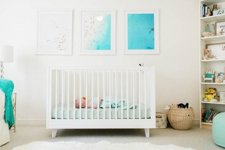 A baby sleeps in a white crib in a nursery with white walls and the best crib mattress. Three framed blue and white prints are hung above the crib, adding a touch of charm. To the right, a basket holds toys and books on neat shelves, completing the room's calm ambiance.