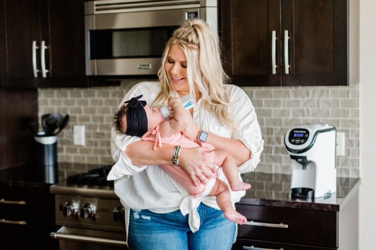 A woman is standing in a kitchen holding a baby. She is feeding the baby a bottle using a Formula Pro Mini. The woman has long blonde hair and is wearing a white shirt and blue jeans. The baby is dressed in a pink outfit with a black headband. There is a coffee machine on the counter.
