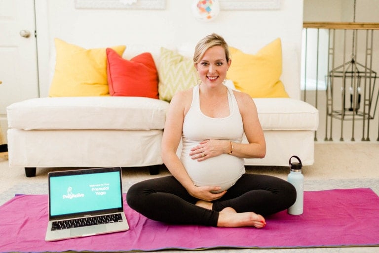 A PregActive woman sits cross-legged on a pink yoga mat, holding her belly with a gentle smile. Behind her is a white couch adorned with colorful pillows. A laptop displaying "Prenatal Yoga" and a water bottle are placed on the mat beside her, encapsulating serene preparation.