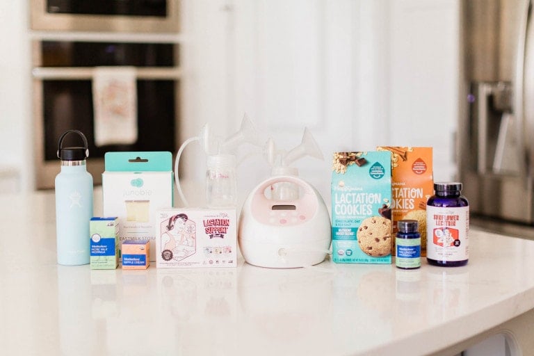 A kitchen countertop displays various breastfeeding and postpartum products that increase milk supply, including a water bottle, breast pump, nipple cream, lactation cookies, supplements, and breast milk storage bags. An oven and refrigerator are visible in the background.
