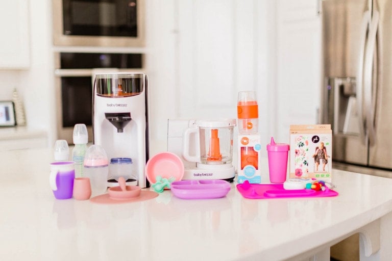 A collection of products for feeding baby is displayed on a kitchen counter, including a bottle warmer, baby food maker, various bottles, sippy cups, bibs, a plate with utensils, pacifiers, and a teething ring. The background showcases kitchen cabinets and a refrigerator.