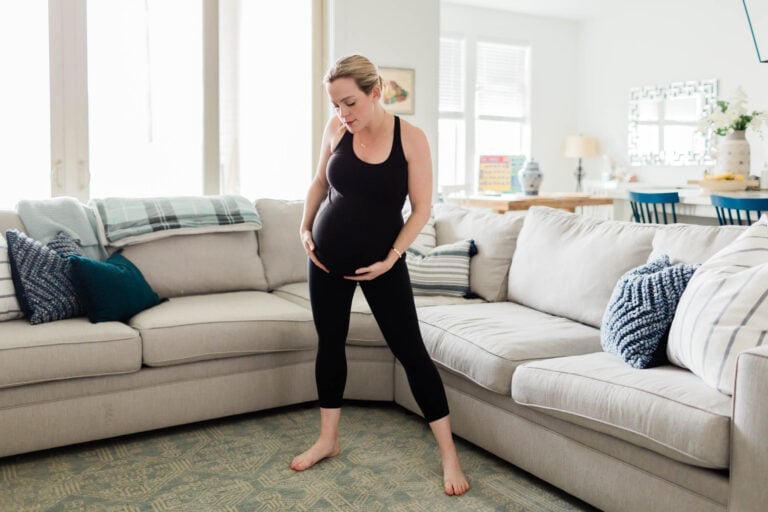 A pregnant woman in a black tank top and leggings stands between two beige sofas inside a bright living room. Holding her belly and looking downward with a neutral expression, she seems to be contemplating signs that labor is starting. The room is decorated with various cushions, a blanket, and visible furniture.