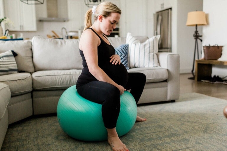A pregnant person in a black tank top and leggings is sitting on a teal birthing ball in a living room. They are holding their belly with both hands. The room has a light-colored couch, pillows, a table lamp, and a tote bag in the background.
