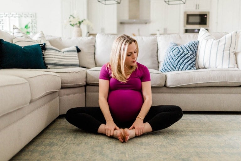 A pregnant woman is sitting on the floor in a butterfly position, performing hip opening stretches by holding her feet with her hands. She is wearing a purple top and black leggings. Behind her, there is a light-colored sectional sofa with blue and white pillows, creating a cozy living room scene.