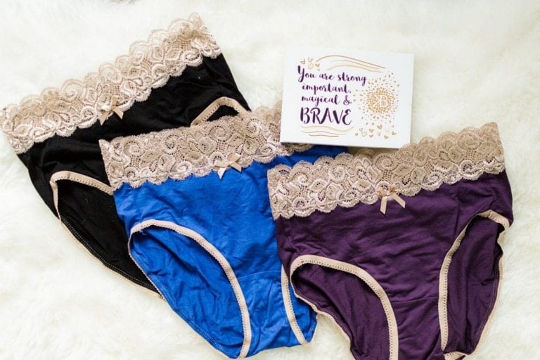 Three pairs of women's Kindred Bravely postpartum underwear in black, blue, and purple with beige lace trim are laid out on a white, furry surface. A card with the text "You are strong, important, magical, & BRAVE" is placed above the underwear.