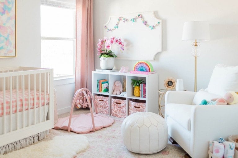 The Palm Beach Nursery features a pastel-themed decor with a white crib, pink and white checkered bedding, a storage unit with various items, a pink play gym, a white armchair, and a white lamp. Pink curtains frame the window adorned with decorative flowers, books, and a rainbow.