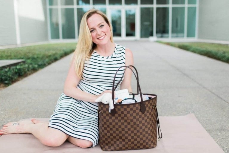 A woman with long blonde hair, wearing a striped dress, sits on a mat outdoors. She has a large checkered handbag next to her, filled with various items—a perfect illustration of what to pack in your diaper bag. The background features a modern building with glass doors and windows.
