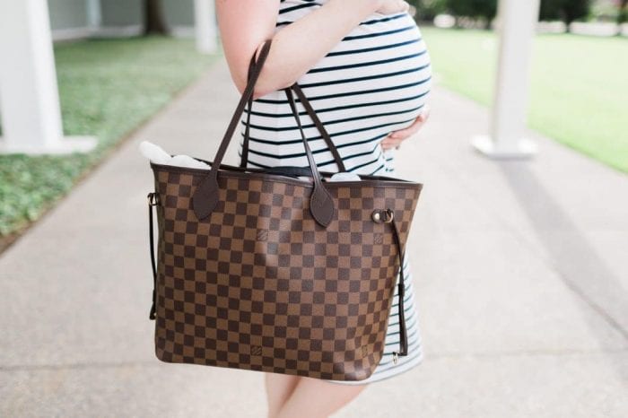 A person in a blue and white striped dress holds their pregnant belly with one hand and a brown checkered handbag, packed with essential hospital bag items, with the other. The background features a walkway and green lawn with columns in view.