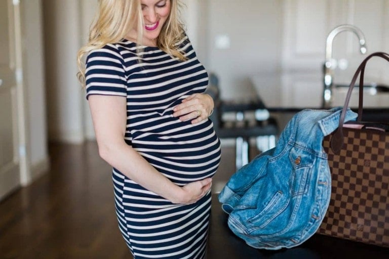 A pregnant woman wearing a black and white striped dress, a maternity clothes essential, holds her belly and smiles. She is standing near a counter with a checkered handbag and a denim jacket hanging on it. The background features a kitchen with hardwood flooring.