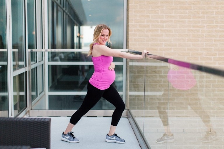 A pregnant woman in a pink tank top and black leggings is doing a leg stretch on a balcony, aiming to boost energy during pregnancy. She is holding onto the glass railing for support and smiling at the camera. The building behind her has large windows and a brick wall.