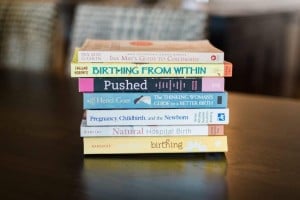 A stack of pregnancy books and parenting guides on a dark wooden table. Titles include "Ina May's Guide to Childbirth," "Birthing from Within," "Pushed," "The Thinking Woman's Guide to a Better Birth," "Pregnancy, Childbirth, and the Newborn," and "Mindful Birthing.