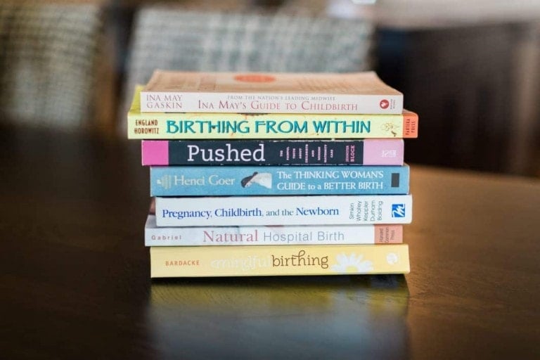 A stack of pregnancy books and parenting guides on a dark wooden table. Titles include "Ina May's Guide to Childbirth," "Birthing from Within," "Pushed," "The Thinking Woman's Guide to a Better Birth," "Pregnancy, Childbirth, and the Newborn," and "Mindful Birthing.