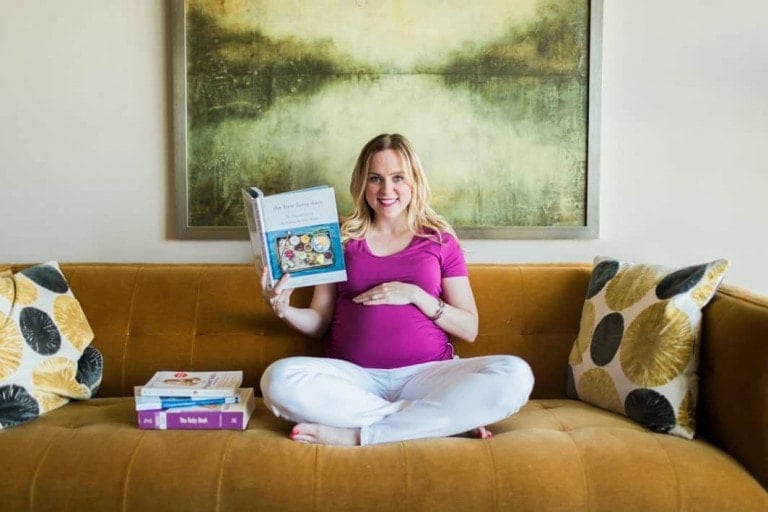 A pregnant woman in a purple shirt and white pants sits cross-legged on a mustard-colored sofa, holding a parenting book. There are two patterned pillows on the sofa and a few other motherhood books beside her. A large abstract painting is mounted on the wall behind her.