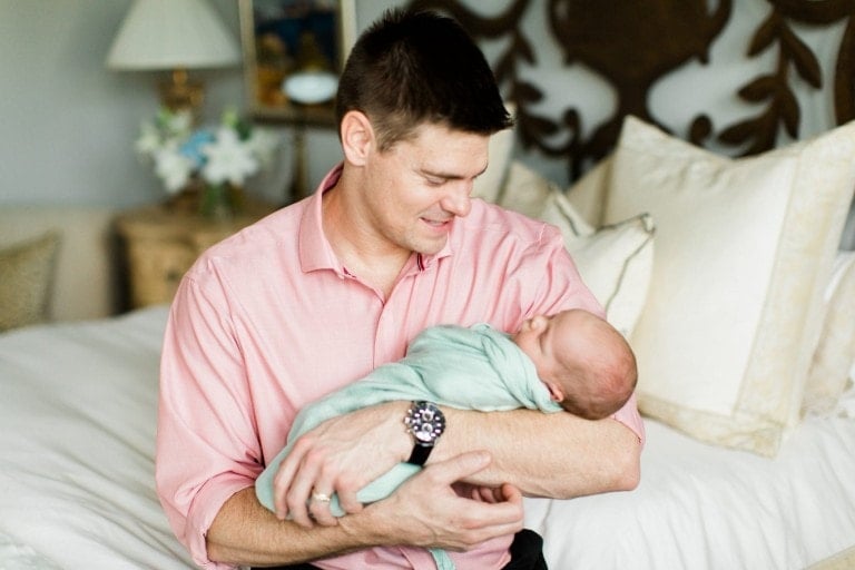 A man in a pink shirt is sitting on a bed and smiling at a newborn baby wrapped in a light green blanket that he is holding in his arms, fully embracing the truth that sons need their fathers. The room has a patterned headboard, cream-colored pillows, and a bedside table with a lamp and flowers.