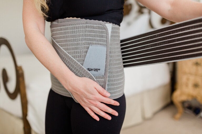 A person wearing a black shirt and black pants is adjusting a grey Mamaway abdominal binder around their waist. The background includes a bed with white bedding and partially visible beige furniture.