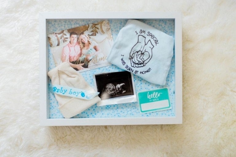 A baby shadow box contains a photo of a couple, a baby onesie with "I am special, I was born at home!" printed on it, a small hat labeled "Baby boy," an ultrasound image, and a "hello my name is" name tag on a white, fluffy surface.