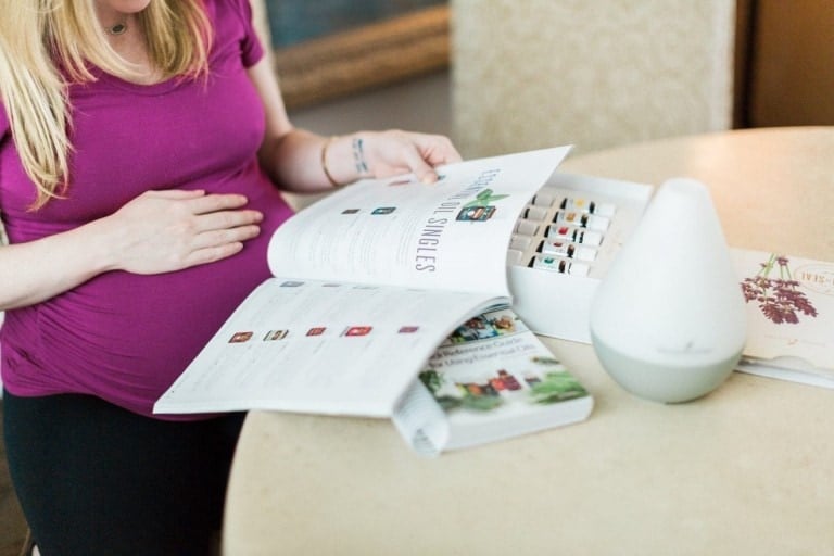 A pregnant woman in a magenta shirt is looking at a catalog labeled "ESSENTIAL SINGLES" on a table. Next to her, there is an aroma diffuser, a box of essential oils for labor, and other booklets. She rests one hand on her belly and holds the catalog with the other.