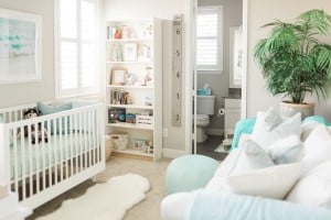 An ocean-inspired nursery features a white crib with light green bedding, a shelving unit filled with books and toys, and a growth chart on the doorframe. A toilet and sink are visible through an open door. A sofa with pillows and a large plant complete the serene space.