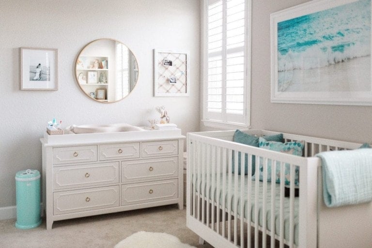 Design a nursery featuring a white crib with teal bedding, a white dresser with a changing pad, a round mirror, wall decor including frames and pictures, a teal diaper pail, and a window with white shutters. The nursery has light walls and carpeting for an airy feel.