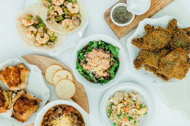 A spread of 30 minute dinner ideas includes shrimp tacos, fried chicken, grilled chicken thighs, crackers with cheese, a bowl of salad, a bowl of rice with vegetables, and a bowl of chili topped with shredded cheese. A salt and pepper shaker is present on a cutting board.