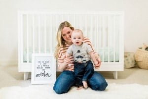 A woman with blonde hair sits on a white rug in front of a white crib, holding a baby in her lap. The baby, wearing a gray shirt and jeans, has been given a lovely fall name. Next to them is a sign that reads, "I'm so glad I live in a world where there are Octobers.