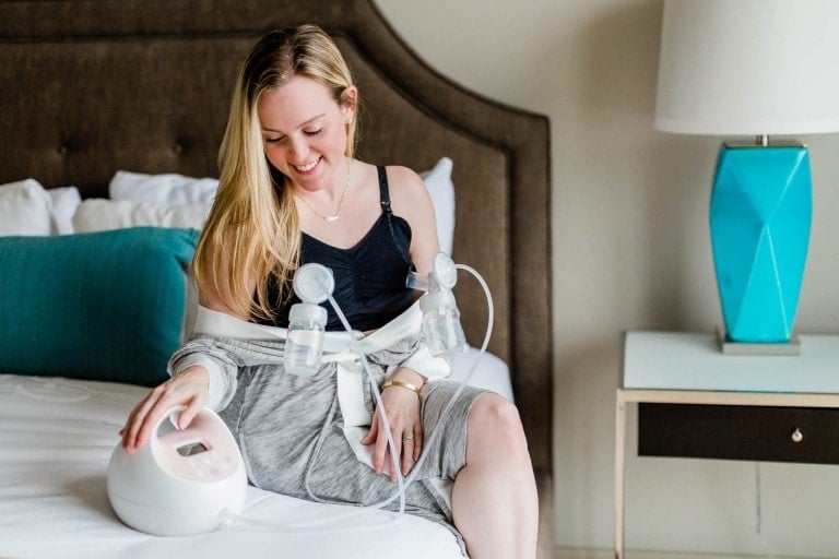 A blonde woman is sitting on a bed, exclusively pumping with a breast pump. She is wearing a black top and a gray skirt, with the device attached to her top. Smiling, she looks down at the pump placed beside her on the bed. A lamp sits on a nightstand nearby.