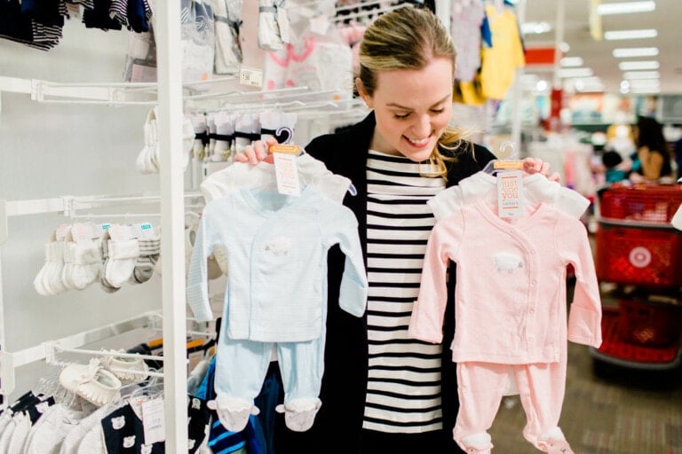 A woman in a store is holding up two baby outfits, one blue and one pink, and smiling. She is standing in front of a display with more baby clothing and socks. A red shopping cart is visible in the background, making her Target baby registry experience even more delightful.