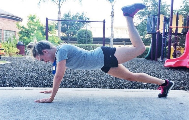 A woman wearing a grey T-shirt and black shorts is in a plank position with one leg raised, focusing on a butt workout. She is exercising on a concrete surface in an outdoor playground area with trees and playground equipment in the background.