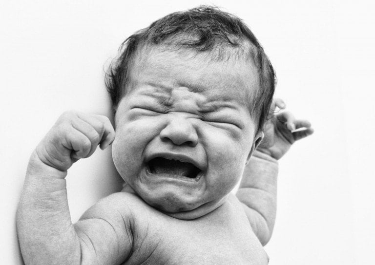 Black and white close-up of a crying newborn baby. The baby has furrowed brows, closed eyes, and an open mouth. Both arms are raised with clenched fists, showing clear distress. This raw moment captures the urgency to soothe a fussy baby against the backdrop of plain white.