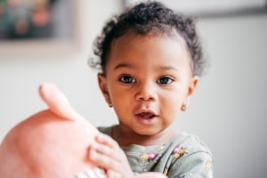 A toddler with curly hair, wearing a green top with floral patterns, looks directly at the camera. They are holding a pink stuffed toy with both hands. The background is blurred, showing part of a wall and some artwork — perhaps perfect inspiration for girl names that start with "A.