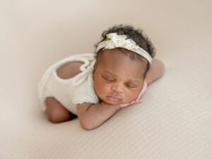 A newborn baby dressed in a white, knit onesie and a floral headband is sleeping on a soft beige blanket. The baby, perhaps destined for one of those beautiful girl names that start with Z, has their arms tucked under their chin and is lying on their stomach.
