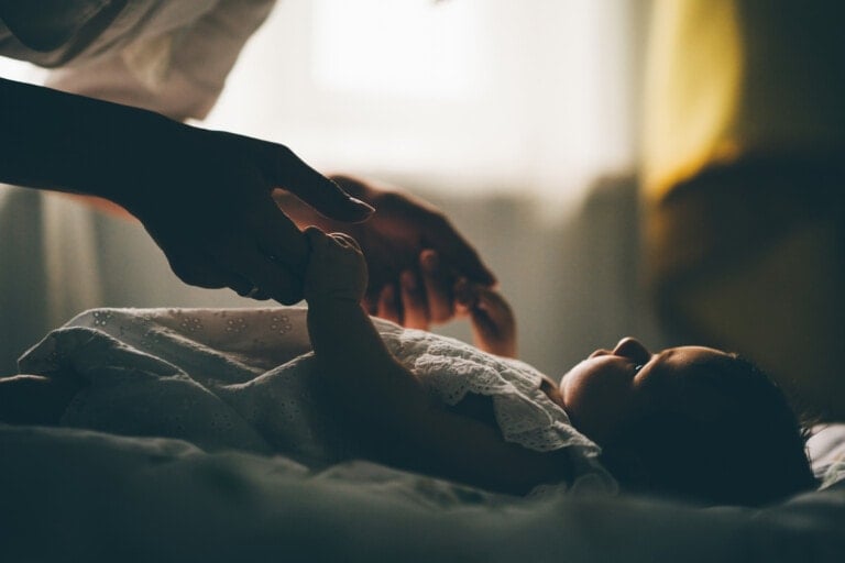 A baby lies on its back in a dimly lit room, wearing a white outfit. Two adult hands reach towards the baby from the left and right side of the frame, gently holding the baby's hands as it begins to self-soothe. The background is softly blurred with light coming through a window.