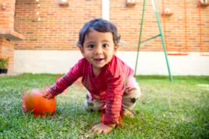A toddler wearing a red long-sleeve shirt and patterned pants is on the grass with one hand touching an orange ball. The child, possibly one of those unique boy names that start with Y, is smiling and positioned in front of a brick wall and a swing set.
